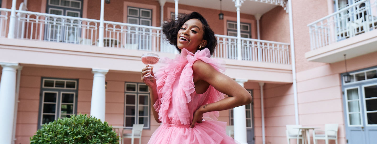 African woman in a pink tulle dress in front of a pink building, thabsie, brutal fruit, the suite, the suite edit, belmond mount nelson hotel, south africa, pink dress, woman with drink, woman with champagne